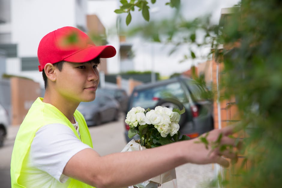 A young delivery person wearing a red cap, white t-shirt, and high-visibility yellow vest is placing a floral bouquet against a bush on a residential street. The bouquet features white roses and hydra