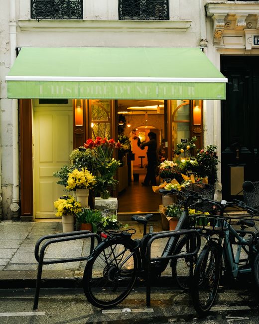 A floral shop with a green awning displaying white text, open to reveal an interior filled with colorful flower arrangements and bouquets. The shop's entrance is decorated with a variety of fresh flow
