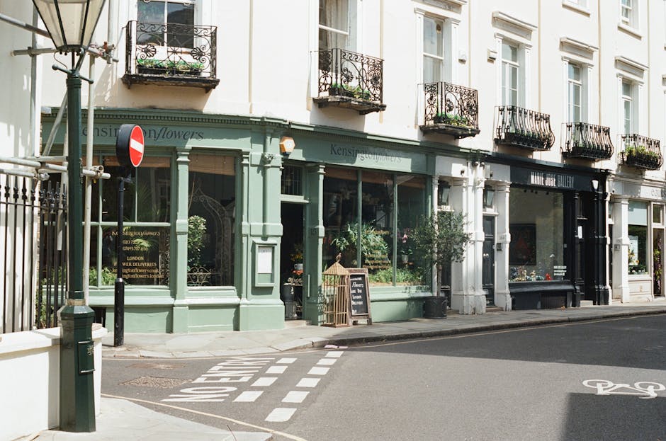 Exterior view of a flower shop with a light green facade, large display windows filled with various flower arrangements, and potted plants near the entrance. The shop is situated on a street corner, w