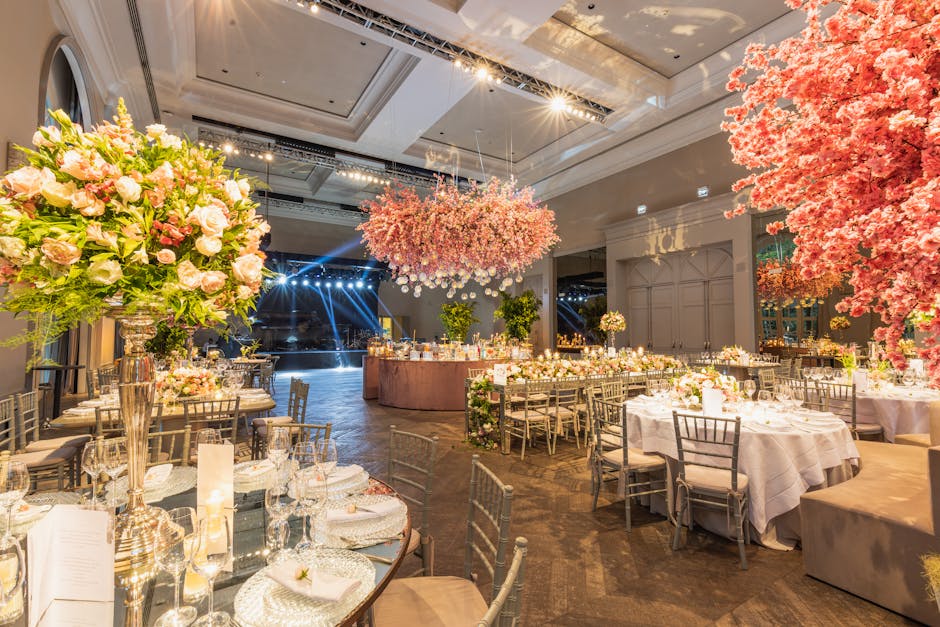 An elegantly decorated indoor event space featuring floral arrangements and table settings for a wedding reception. In the foreground, there is a large bouquet of pale pink roses, white peonies, and g