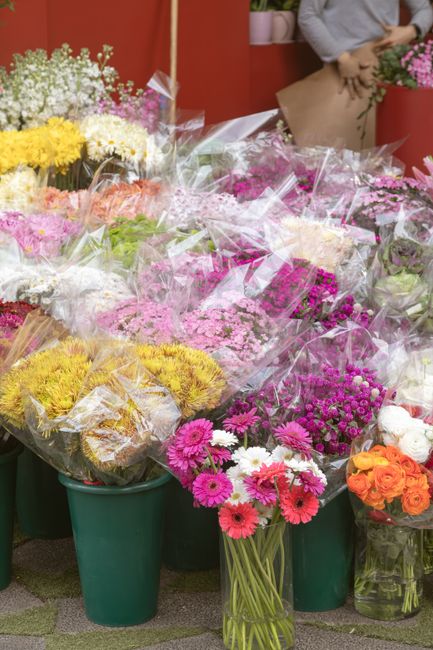 A vibrant display of fresh flowers at a florist shop, featuring an assortment of colorful blooms including pink gerberas, white daisies, and orange roses arranged in clear plastic wrapping. The flower