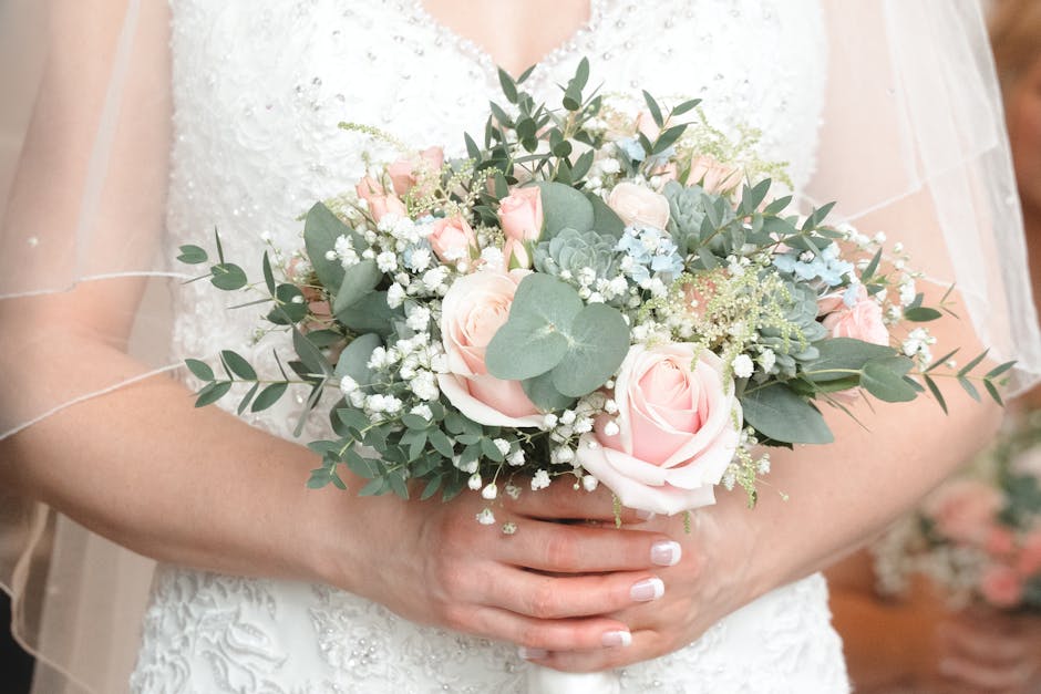 A person wearing a white wedding dress with lace details is holding a floral bouquet composed of soft pink roses, white baby's breath, and green eucalyptus leaves. The arrangement is rounded and lush,