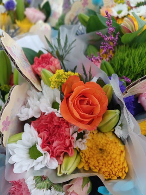 A close-up view of a floral bouquet featuring vibrant orange roses, white daisies with green centers, pink carnations, yellow chrysanthemums, and sprigs of purple lavender. The flowers are wrapped in 